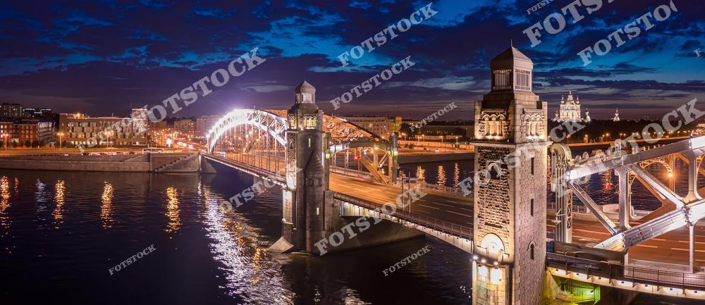 На фотографии изображён Большеохтинский мост, освещённый ночной иллюминацией. Этот мост, один из символов Санкт-Петербурга, соединяет берега реки Невы. На фоне видны исторические здания и спокойная водная гладь, отражающая огни. Ночное освещение подчёркивает красоту конструкции и придаёт мосту особое очарование.